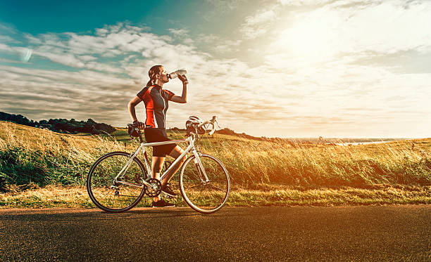 Female race cyclist stands next to bike near the sea Full lenght portrait of female cyclist, who takes a break and drinks water. The sun sets and the woman wears sports clothes, but has removed her bike helmet. It is mounted on the bike, while she is resting and enjoying the water after she has been racing on her bike. racing bicycle photos stock pictures, royalty-free photos & images