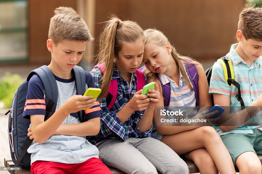 elementary school students with smartphones primary education, friendship, childhood, technology and people concept - group of happy elementary school students with smartphones and backpacks sitting on bench outdoors Child Stock Photo