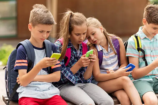 primary education, friendship, childhood, technology and people concept - group of happy elementary school students with smartphones and backpacks sitting on bench outdoors