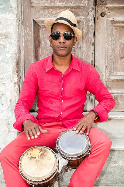 A well known professional Cuban conga drum player percussionist and the afro-cuban Santeria priest in red clothes and Panama hat sitting in front of an old door, playing bongo drums, Old Havana, Cuba.