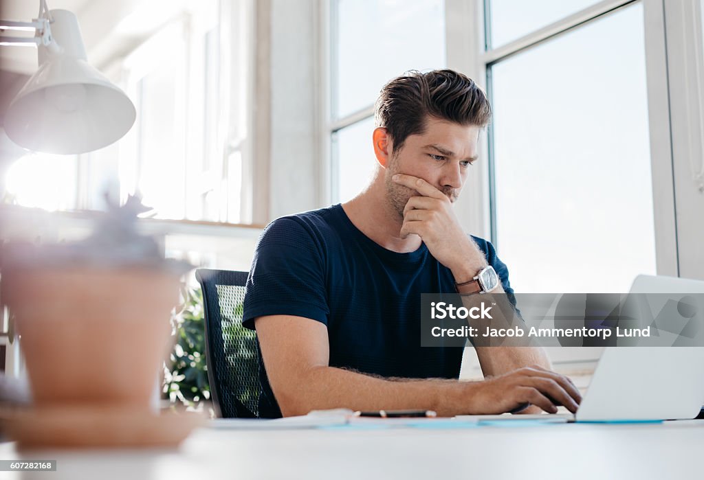 Pensive young male executive using laptop at his desk Young man working on laptop in office and thinking. Young male executive using laptop at his desk. Concentration Stock Photo