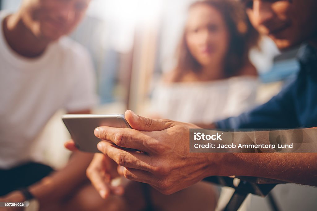 Young man showing mobile phone to friends Young friends sitting outdoors and looking at smartphone. Young man showing something interesting to friends. Focus on mobile phone in hand. Friendship Stock Photo