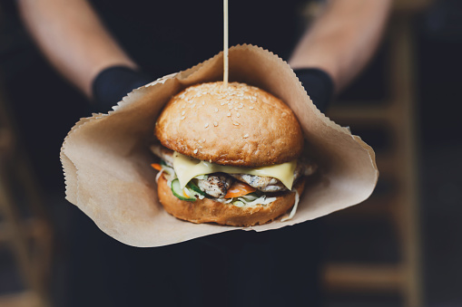 Fresh burger cooked at barbecue outdoors in craft paper. Cookout american bbq food. Big hamburger with steak meat and vegetables closeup with chef unfocused at background. Street food, fast food.