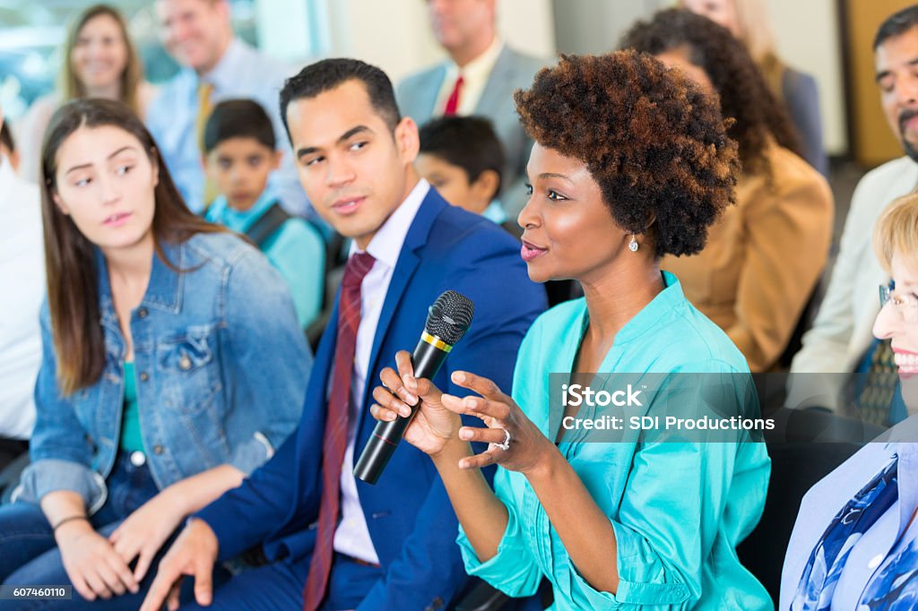 Confident African American woman asks question during a meeting Beautiful  mature African American woman gestures while asking a political candidate a question during a town hall meeting. A diverse group of people are sitting with her in the audience. Town Hall Meeting Stock Photo