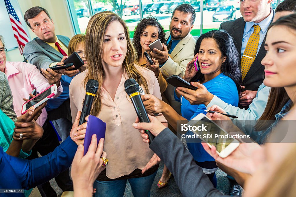 Mid adult Hispanic female politician answers questions after her speech Confident mid adult female political candidate pauses to take questions from the press after her campaign speech. A large group of reporters are gathered around her with smart phones and microphones. Politician Stock Photo