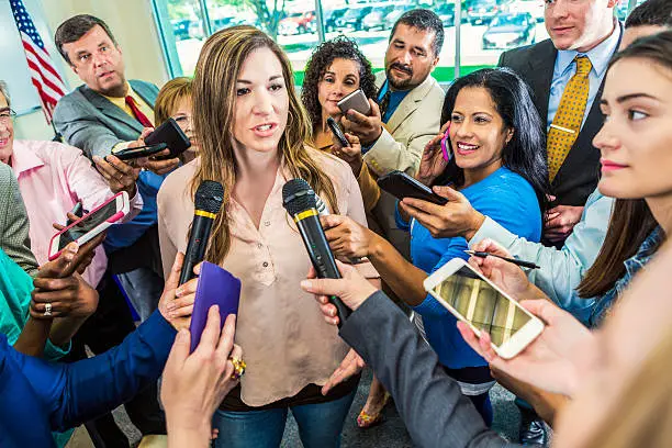 Confident mid adult female political candidate pauses to take questions from the press after her campaign speech. A large group of reporters are gathered around her with smart phones and microphones.