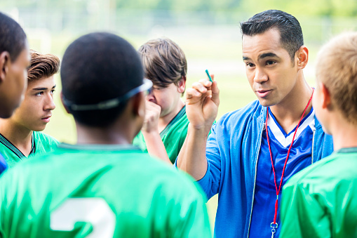 Serious mid adult male Hispanic soccer coach gestures while making a point during time out. the players are watching their coach. The players are wearing green jerseys and the coach is wearing a blue jersey.