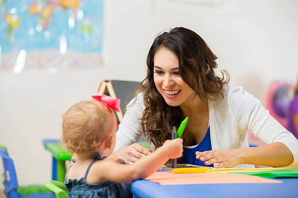 Pretty daycare teacher helps toddler with coloring project Beautiful Hispanic female daycare teacher leans on table and helps cute blonde toddler girl with school project. Little toddler girl is holding crayons and the teacher is smiling at preschooler. early morning stock pictures, royalty-free photos & images