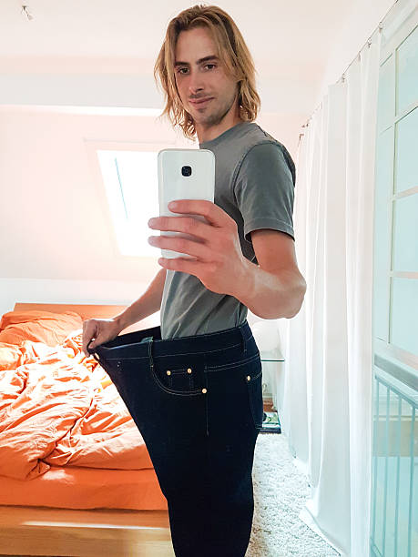 istock Young man taking selfie in mirror showing weight loss 607476688