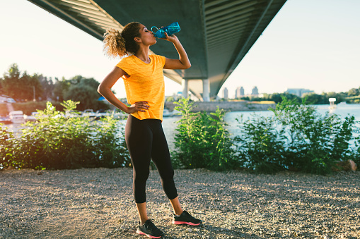 Latina Runner running in the city near river. Standing under the bridge, making break to drink some water. Cityscape in background.