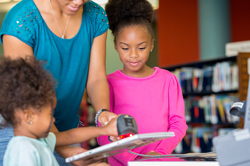 African American family scanning books at their local library. Cute elementary age African American girl, adorable African American toddler girl and their mid adult African American mother standing at a desk scanning books at the local library.
