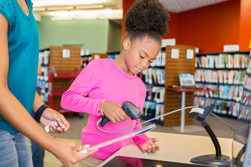 Elementary age African American girl stands at a desk with her mother at the local library and scans a book. Cute elementary age African American girl with her mid adult African American mother stand at a desk together as the girl scans books in the library.