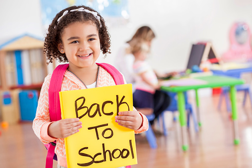 Cheerful little girl on the first day of school holds a 'Back to School' sign on the first day of preschool. She has curly black hair and is wearing a pink patterned sweater and a pink backpack. A teacher works with a student in the background.