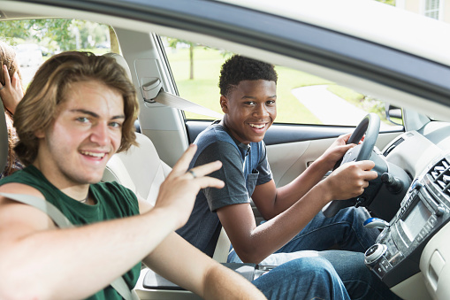 An African American teenage boy in the driver's seat of a car. He and his friend in the passenger seat are looking out the window at the camera.
