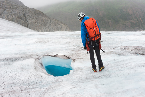 Man looks curiously into a blue-colored moulin hole in ice that could be hundreds of feet deep on a trekking adventure on Lemon Glacier, Juneau Icefield, Juneau, Alaska, USA