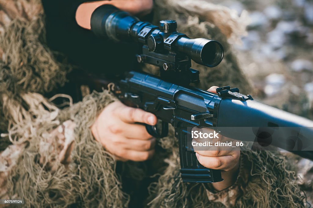 Soldier in camouflage with a sniper rifle. The soldier disguised in a balaclava and military camouflage aims at the sight of a sniper rifle.  War Zone. Aggression Stock Photo