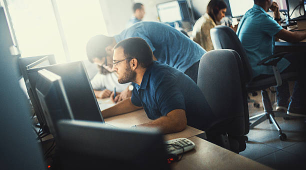 Software developers at work. Group of mid 20's and 30's men and women engaged in application development job. They are seated by long desks back to back, each person in front of dual screen computer. Bad posture is innevitable. Back lit, toned image. bad posture stock pictures, royalty-free photos & images