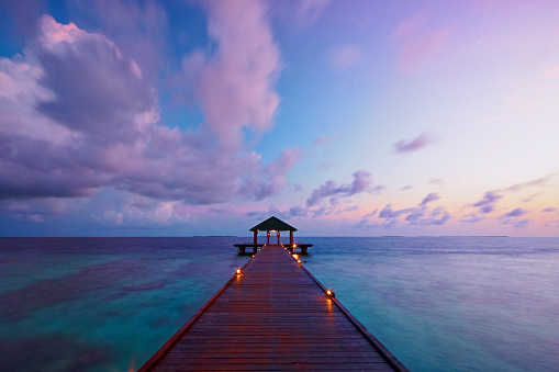 Dawn in the Maldives. A pier juts out into the tropical waters around the island of Vilamendhoo whilst, overhead, the sun turns the sky to spectacular reds & pinks. Various corals can be seen in the clear waters below.