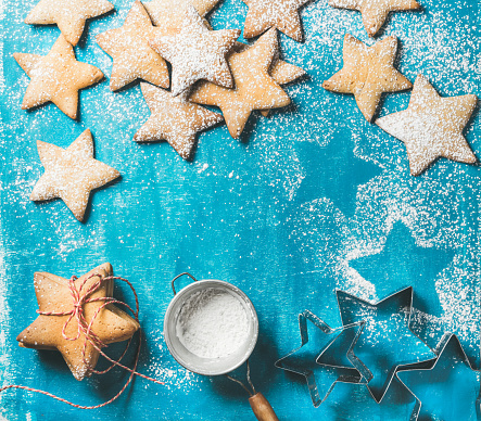Christmas or New Year holiday food background. Sweet gingerbread cookies in shape of star with sugar powder in sieve and metal shapes on bright blue painted plywood background, top view, copy space