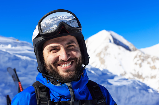 Smiling young man hiking on ski resort. With ski wear, goggles and helmet, backpack. Looking at camera. On background snow mountain peak. Focus on foreground.