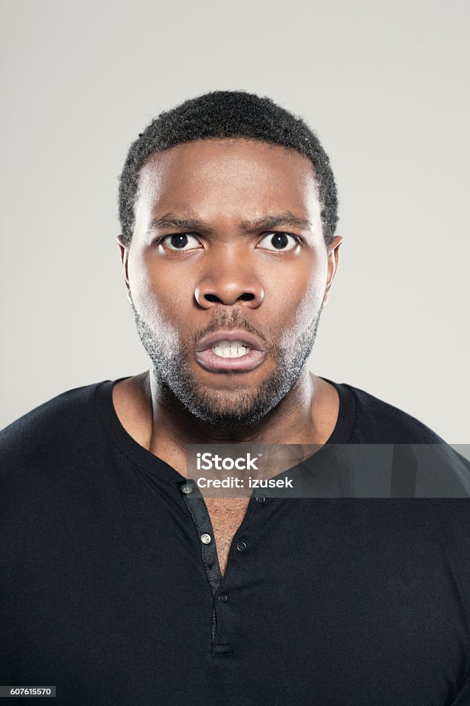 Portrait of angry afro american young man Portrait of angry afro american young man wearing black t-shirt, standing against grey background, clenching teeth, staring at camera. Adult Stock Photo