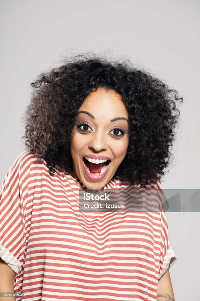 Surprised afro american woman laughing Portrait of surprised afro american young woman wearing striped top, standing against grey background and shouting at camera. Cheesy Grin Stock Photo