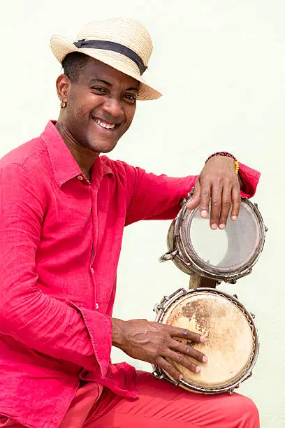 A well known professional Cuban conga drum player percussionist and the afro-cuban Santeria priest in red clothes and Panama hat standing against a green wall, holding bongo drums, Old Havana, Cuba.