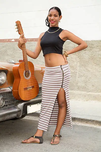 Beautiful young Cuban woman with a guitar, standing on the street by an old car, smiling, looking at camera, Havana, Cuba