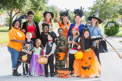 A large, multi-ethnic group of mothers and children at halloween. The boys and girls are wearing various costumes, standing in front of the parents.