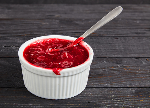 Delicious homemade cranberry sauce in a white bowl with a dessert spoon in it. Berry confiture on dark wooden table.Perfect traditional sweetness for the cup of tea. Close up shot