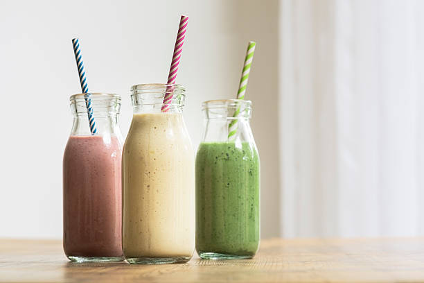 Three glass bottles of freshly blended fruit and vegetable smoothies with a stripy straw in each bottle. The yellow is banana, green is spinach and avocado and pink is strawberry. They are on a wooden table.