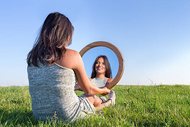 Woman in nature viewing her mirror image Dutch woman sitting on grass looking at  her mirror image. Wooden mirror on  woman's knee with reflection of herself. The european woman is sitting in the green meadow in a natural rural landscape. I choose a lot of blue sky as background. On this sunny day in summer season the female model is enjoying the warm sunny weather. Concept of beauty, feminine, women, pretty, black hair, fun, enjoy, pleasure, happy, happiness, pasture, laugh, laughing, enjoy, enjoying. behavior femininity outdoors horizontal stock pictures, royalty-free photos & images