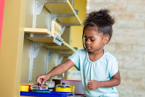 Young African American preschool girl playing with a toy kitchen. Cute little girl grabbing toy food items and fake crockery while playing with a pretend kitchen.