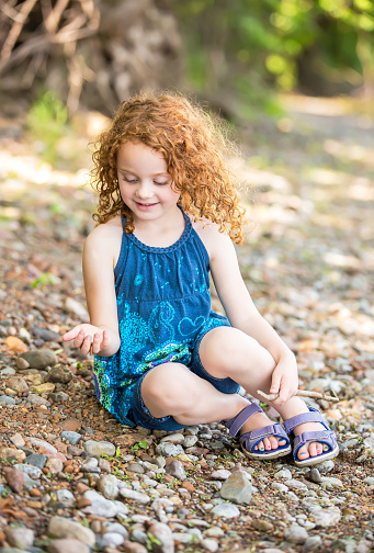 Young girl with curly red hair sitting on the bank of the Mississippi River on a summer day in Minnesota, USA. She is looking down at the rocks she has just picked up with her right hand.