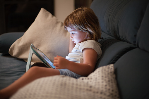 blonde three years old baby shirt and shorts, sitting comfortably in sofa inside home at night reading and watching digital tablet, face illuminated by the light of the screen