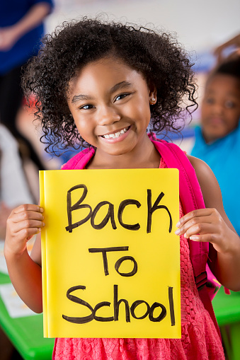 Excited girl is happy about the first day of school. She is holding a yellow 'Back to School' sign. She has curly black hair. Students are in the background.