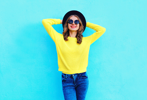 Fashion happy pretty smiling woman wearing a black hat and yellow knitted sweater over colorful blue background
