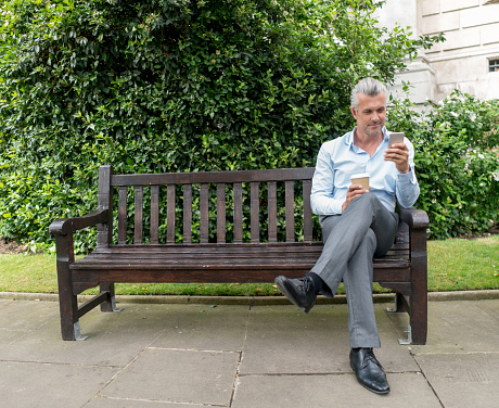 Business man on his coffee break at the park texting on his mobile phone and looking happy