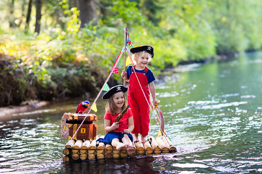 Kids dressed in pirate costumes and hats with treasure chest, spyglasses, and swords playing on wooden raft sailing in a river on hot summer day. Pirates role game for children. Water fun for family.