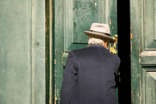 Rome, Italy - March 2, 2008: A senior man nattily dressed in a fedora and mohair coat pushes open an old green distressed sunlit door. 