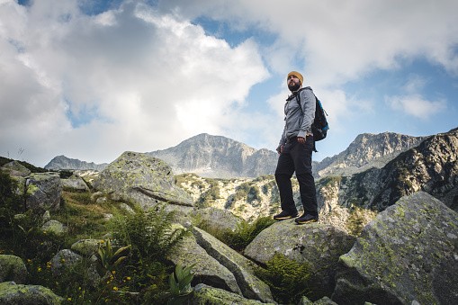 Traveler with backpack stands on a huge rocks and looking around. Blue sky over mountain range on background.