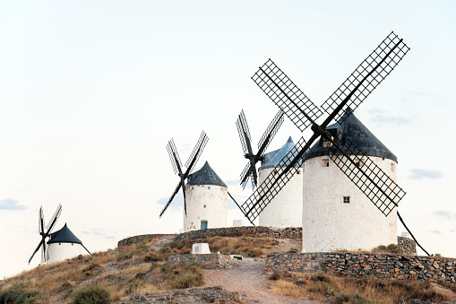 Consuegra, Toledo, Spain: A row of four traditional Spanish windmills on a hill, contrasting against an almost white sky with few clouds just after sunset.