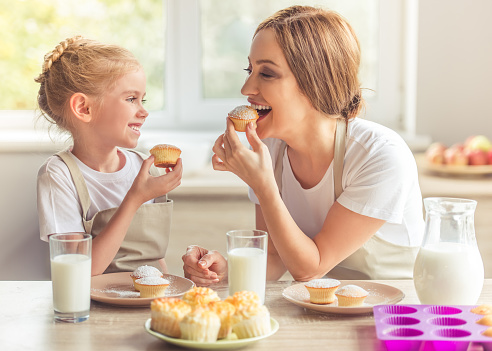 Beautiful woman and her cute little daughter in aprons are looking at each other and smiling while tasting their muffins