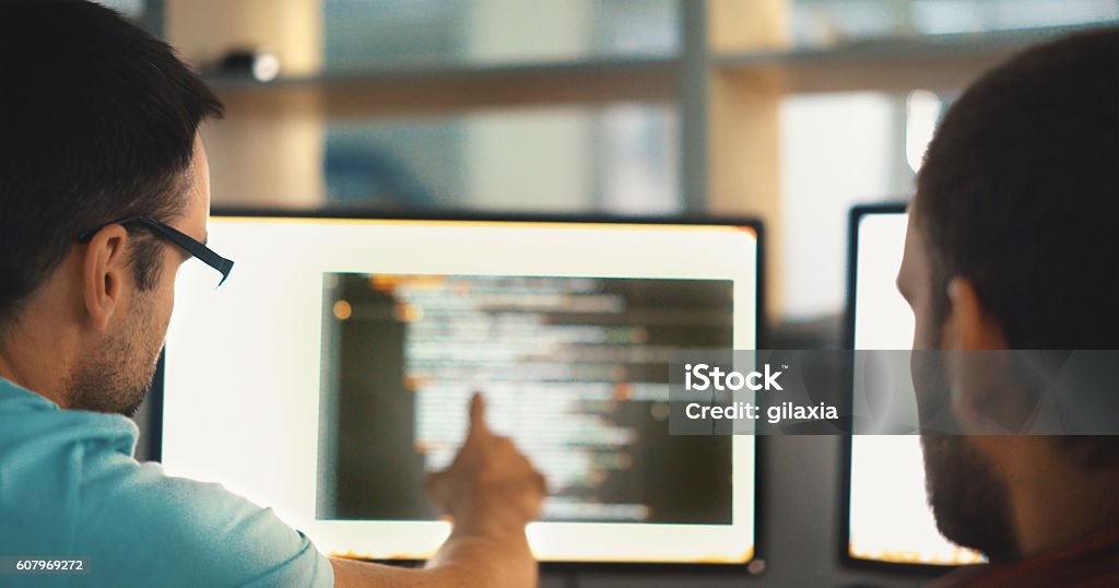 Software developers. Rear view of two partially unrecognizable men sitting in front of a computer. One of them is pointing at a screen where are several lines of computer code. Computer Software Stock Photo