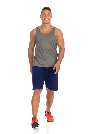 Young man in blue shorts, gray tank top and orange sneakers, walking and looking at camera. Front view. Full length studio shot isolated on white.