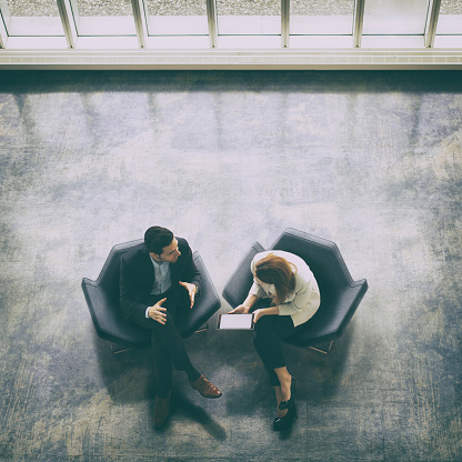 A high-angle view of a businessman and a businesswoman sitting in the office building lobby and using a digital tablet