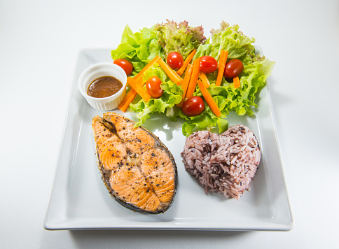 clean eating grill salmon steak with rice berry, red oak, green oak, carrot, tomatoes and dressing with sesame soy sauce in a white ceramic dish with a white background