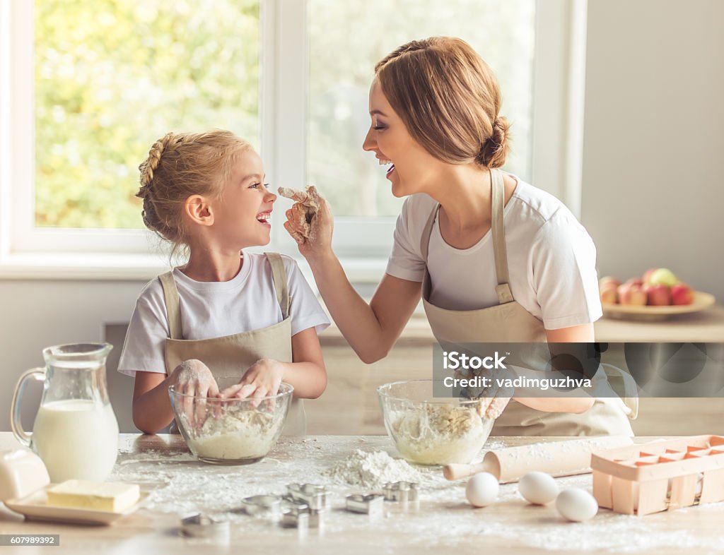 Mother and daughter baking Cute little girl and her beautiful mom in aprons are playing and laughing while kneading the dough in the kitchen Cooking Stock Photo