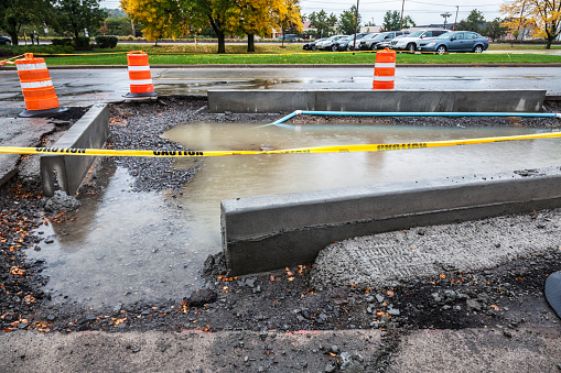 New, reconfigured concrete curbs and entrance lanes are under construction on a messy, rainy day in a suburban retail shopping center asphalt surface parking lot. The incomplete road construction zone excavated section is filled with a large puddle of rainwater, and is cordoned off with yellow CAUTION tape, and surrounded and protected by bright orange striped traffic cone barrels.