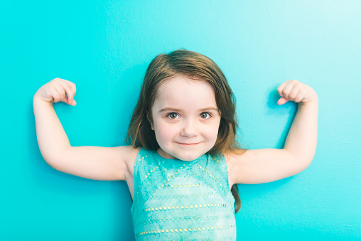 This is a horizontal, color photograph of an adorable four year old wearing a dress standing against an aqua wall as she looks at the camera as she raises her arms showing off her little bicep muscles demonstrating how strong she is. Photographed with a Nikon D800 DSLR camera.